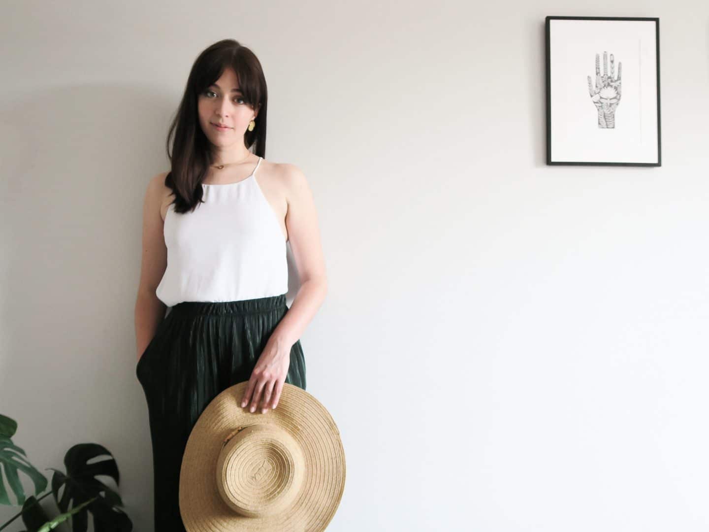 Besma in a white strappy top, green wide-leg trousers holding a straw hat